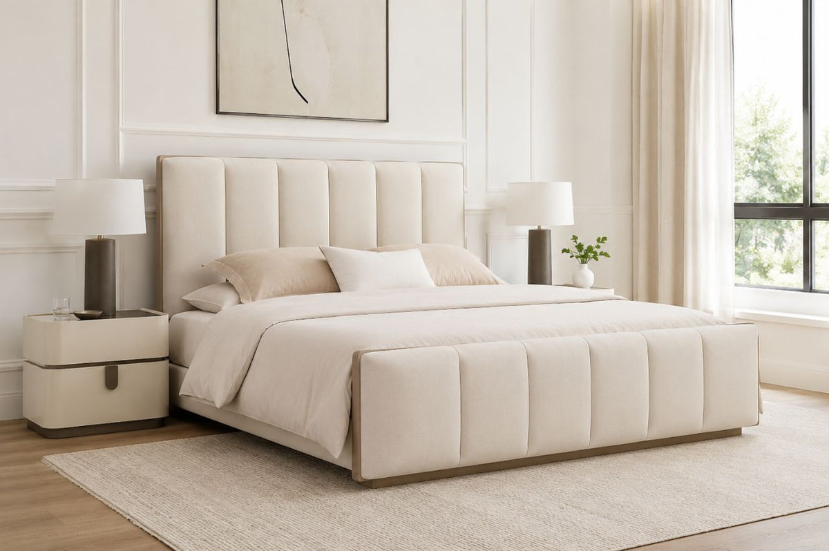 Modern upholstered cream bed with a channel-tufted headboard and matching footboard in a bright minimalist bedroom featuring bedside tables, lamps, and large windows.